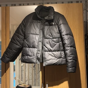 Svart pufferjacka från The North Face - Säljer en svart pufferjacka från The North Face med hög krage och klassisk logga på bröstet och ryggen. Jackan har quiltad design, dragkedja framtill och är perfekt för kalla dagar. Snygg och enkel att matcha till alla outfits.