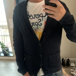 Mörkblå kofta med knappar från Dressmann - Säljer en stilren mörkblå kofta med knappar framtill och två praktiska fickor. Koftan har långärmad design och är tillverkad i ett mjukt, ribbat material som ger en snygg struktur. Perfekt för lager-på-lager och passar till jeans eller chinos. Mitt pris 549kr