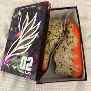 Puma MB.01 M.E.L.O. sneakers camo/orange - Säljer ett par Puma MB.01 Digital Camo M.E.L.O. sneakers med beige och brunt camo-mönster och neonorange sula. Skorna har snörning, högt skaft och coola detaljer på ovandelen. Perfekta för dig som vill sticka ut med en unik och sportig look.