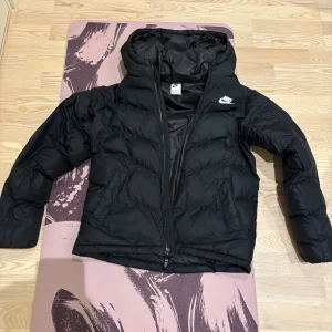 Svart pufferjacka från Nike XL (barn) - Svart pufferjacka från Nike i storlek XL. Jackan har huva, dragkedja framtill och två sidofickor. Klassisk Nike-logga på bröstet och quiltad design. Perfekt för kalla dagar när du vill hålla stilen och värmen.