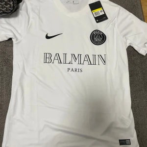 Vit PSG x Balmain fotbollströja Nike - Snygg vit fotbollströja från Nike i samarbete med PSG och Balmain. Tröjan har korta ärmar, svart Nike-logga och PSG-emblem på bröstet samt Balmain Paris-tryck framtill. Tillverkad i lätt och ventilerande material, perfekt för match eller träning. Storlek S passar som M också!!