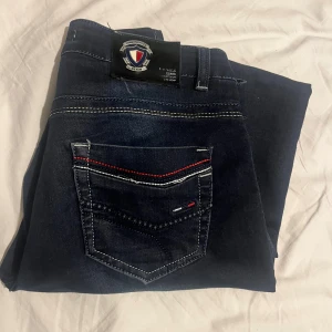 Tommy Hilfiger mörkblå jeans straight - Mörkblå jeans från Tommy Hilfiger vintage vibemed raka ben och klassisk femficksdesign. Snygga kontrastsömmar i gult och rött samt logga på bakfickan och läderpatch i midjan. Jeansen har normal passform.