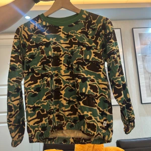 Camouflage tröja från Mini Rodini (140/146) - Säljer en långärmad tröja från Mini Rodini med grön, brun och beige camouflage-mönster. Tröjan har gröna ribbade muddar vid halsen och resår vid ärmslut och nederkant. Perfekt för dig som gillar streetstyle och coola prints. Oanvänd!