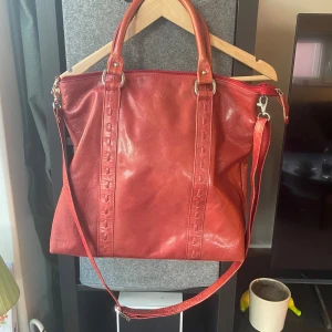 Röd axelväska i skinn från The Monte - Snygg röd axelväska från The Monte i äkta skinn. Väskan har både handtag och justerbar axelrem, dragkedja upptill och flera fack, inklusive ett med dragkedja på baksidan. Perfekt storlek för att få plats med allt du behöver.