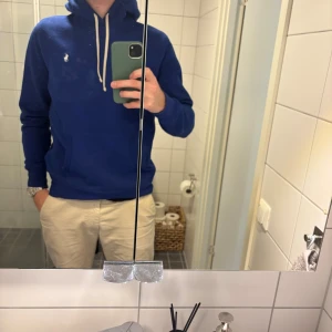 Blå hoodie från Polo Ralph Lauren - Snygg blå hoodie från Polo Ralph Lauren med vit broderad logga på bröstet och vita snören i huvan. Klassisk känguruficka framtill och mjukt material. Bra skick använd fåtalet gånger, storlek M.