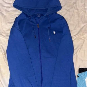 Blå hoodie från Polo Ralph Lauren - Snygg blå hoodie från Polo Ralph Lauren med dragkedja framtill och klassisk vit logga på bröstet. Tröjan har huva med snörning och är tillverkad i mjuk bomull. Perfekt för en chill och stilren look.
