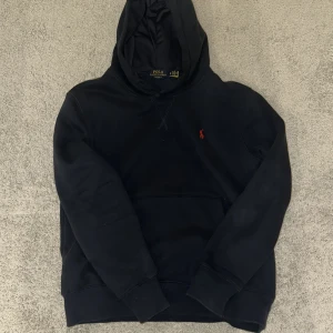 Marinblå hoodie från Polo Ralph Lauren - Marinblå hoodie från Polo Ralph Lauren med klassisk röd logga broderad på bröstet. Tröjan har huva, känguruficka och långa ärmar. Mjuk bomullskvalitet och relaxed fit, perfekt för chill dagar.