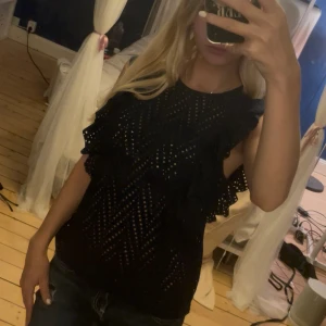 Mörkblå ärmlös blus med volang - En mörkblå ärmlös blus med volangdetaljer längs axlarna. Toppen har ett genombrutet mönster i tyget som ger en cool look och är perfekt för dig som gillar trendiga detaljer. Passformen är normal och materialet känns lätt och luftigt. Från Vero Moda💕
