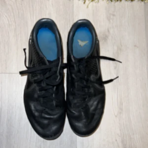 Nike Tiempo svarta fotbollsskor - Svarta Nike Tiempo fotbollsskor med blå detaljer under sulan. Skorna har snörning och är tillverkade i skinn med perforerade partier på sidorna för extra ventilation. Klassisk design med rund tå och låg profil, perfekta för dig som vill ha stilrena och funktionella skor på planen.
