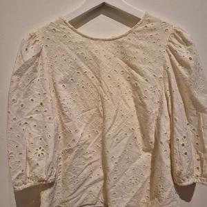 Broderad blus i bomull från Vero Moda - Superfin broderad blus från Vero Moda i offwhite bomull. Blusen har trekvartsärmar med resår vid ärmslut, vackra broderade detaljer och hålmönster över hela plagget. Perfekt för en fräsch och romantisk look.