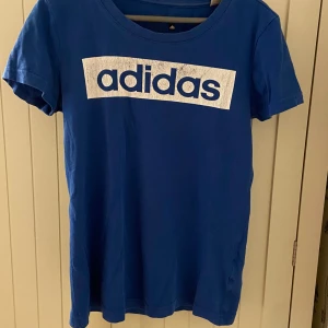 Blå Adidas t-shirt med logga XS - Blå t-shirt från Adidas i modellen Sport Essentials. Stor vit Adidas-logga framtill, rund hals och korta ärmar. Skön passform och tillverkad i mjuk bomull. Perfekt för en sportig och avslappnad look.