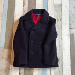 Mörkblå/Röd skeparkavaj jacka från Polo Ralph Lauren - Klassisk mörkblå skepparkavaj från Polo Ralph Lauren med röd innerfoder, dubbelknäppning och bred krage. Jackan har en snygg broderad patch på ärmen med örn och röda detaljer. Perfekt för dig som gillar tidlös och stilren look.