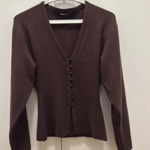 Brun kofta med knappar från Gina Tricot - Snygg brun kofta från Gina Tricot med v-ringning och dekorativa knappar framtill. Modellen har långärmade och är tillverkad i ett mjukt stickat material som känns skönt mot huden. Perfekt att styla med jeans eller kjol för en chill look.