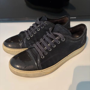 Svarta sneakers från Lanvin i mocka - Snygga svarta sneakers från Lanvin med ovandel i mocka och detaljer i blankt skinn på tå och häl. Skorna har snörning och en beige sula som ger en schysst kontrast. Perfekta för dig som gillar stilrena men ändå unika sneakers.