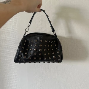 Svart handväska med nitar - Cool svart handväska i skinnimitation med massor av silvriga nitar och studs över hela väskan. Väskan har ett kort handtag med extra nitar,dragkedja fungerar ej. Perfekt accessoar för en edgy look.