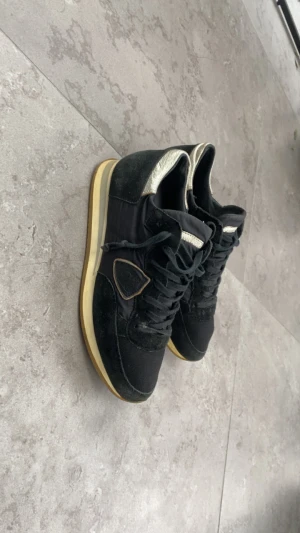 Svarta philippe models - Snygga svarta sneakers från Philippe Model med guldiga detaljer på häl och plös. Skorna har en beige sula, snörning och är tillverkade i en mix av mocka och syntetmaterial. Klassisk rund tå och diskret logga på sidan. Perfekta för dig som gillar stilrena men ändå unika sneakers. Skriv vid minsta fråga🤗