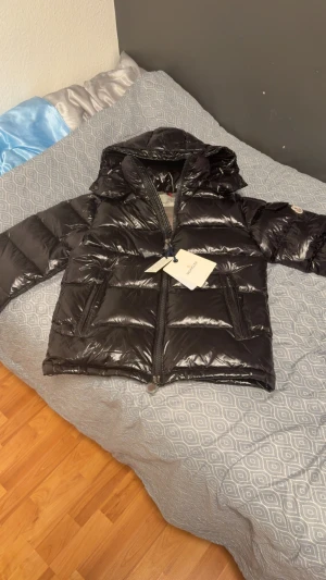 Moncler maya  - Svart pufferjacka från Moncler med glansig finish, huva och två sidofickor med dragkedja. Jackan har Moncler-logga på ärmen och en praktisk ficka med knapp. Perfekt för kalla dagar och har en riktigt clean look.