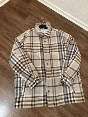 Rutig overshirt från Kappahl, beige/brun - Overshirt från Kappahl i storlek L med rutigt mönster i beige, brunt och vitt. Jackan har knappar framtill, klassisk krage och två sidfickor. Insidan är quiltad för extra värme och komfort. Perfekt för lager-på-lager under höst och vår.