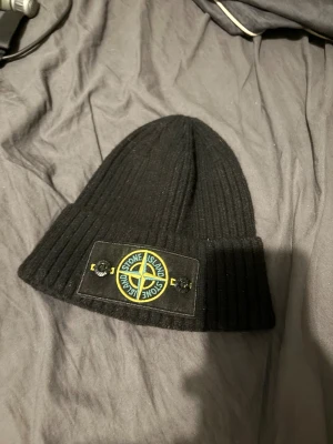 Svart ribbstickad mössa Stone Island - Svart ribbstickad mössa från Stone Island med klassisk patch framtill i svart, gult och grönt. Tillverkad i 100% bomull och har vikbar kant. Snygg logga med knappar och etikett på insidan. Made in Italy.