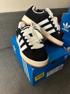 Adidas Campus 00s svart/vit sneakers - Helt nya Adidas Campus 00s sneakers i svart mocka med klassiska vita ränder och vit sula. Skorna har rund tå, platt gummisula och breda vita snören. Perfekt för dig som gillar retrostil och streetwear. Snygg kontrast mellan svart och vitt, med en touch av beige på sulan.