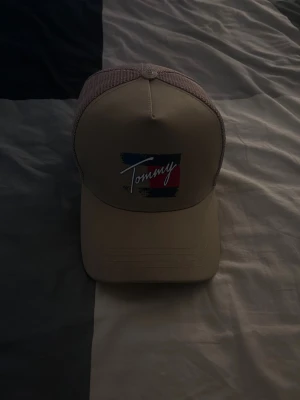 Beige meshkeps från Tommy Hilfiger - Snygg beige keps från Tommy Hilfiger med mesh på sidorna och bak, broderad logga framtill och texten 'Hilfiger' baktill. Justerbar passform och klassisk böjd skärm. Detaljer i rött, vitt och blått på loggan och liten flagga på sidan.