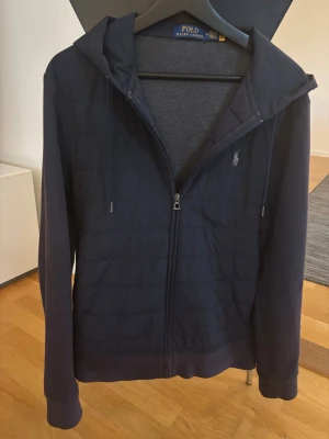 Mörkblå cardigan från Polo Ralph Lauren - Mörkblå cardigan från Polo Ralph Lauren med dragkedja framtill och broderad logga på bröstet. Tröjan har huva, långa ärmar och är fodrad med mjukt grått tyg på insidan. Perfekt för chill dagar och snygg streetstil.