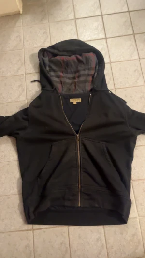 Svart Burberry hoodie med dragkedja - Svart hoodie från Burberry i storlek M med dragkedja och klassiskt rutigt foder i huvan. Två stora fickor framtill och guldfärgad dragkedja. Materialet är mjukt och skönt bomullstyg. Perfekt för dig som gillar stilrena och exklusiva plagg och hoodien har några defekter som ni kan se på bild 3 där det förekommer färgstänk men det går säkert bort med att gnugga med lite sprit men annars riktigt najs hoodie med stor luva! Det är bara att skriva om ni undrar något!☺️☺️☺️🤝👍