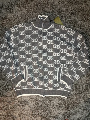 Gucci cardigan  - Säljer en grå stickad kofta från Gucci med hel dragkedja och ribbade muddar. Koftan har ett mönster med vita GG-loggor över hela plagget och vita detaljer vid krage, ärmslut och nederkant. Perfekt för dig som gillar lyxiga streetwear-vibbar.
