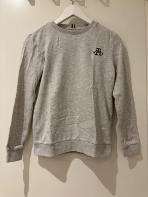 Grå sweatshirt Tommy Hilfiger - Snygg grå sweatshirt från Tommy Hilfiger med diskret logga på bröstet och klassiska ribbade muddar vid ärmslut och nederkant. Tröjan har rund hals och långärmade ärmar, perfekt för chill dagar. Materialet är mjuk bomull som känns skönt mot huden.