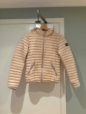Beige pufferjacka från Colmar - Säljer en beige pufferjacka från Colmar med huva och två dragkedjeförsedda fickor. Jackan har quiltad design, lång ärm och Colmar-logga på ärmen. Perfekt för dig som vill ha en lätt och snygg jacka till kyligare dagar.