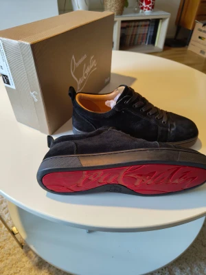 Christian Louboutin svarta sneakers - Svarta sneakers från Christian Louboutin i mocka med klassisk röd sula och rund tå. Skorna har snörning och beige innersula i skinn. Snygg och stilren design med exklusiv känsla, perfekt för dig som gillar lyxiga detaljer.