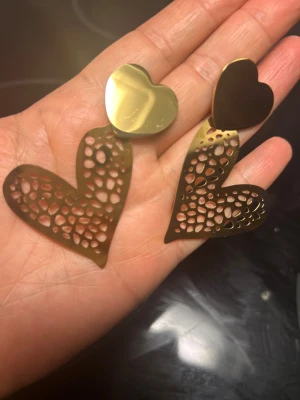 Stål Guldiga hjärtformade örhängen - Säljer ett par örhängen i stål  med hjärtformad design. Örhängena är guldiga och har en cool, perforerad struktur på den nedre delen av hjärtat. Perfekt för dig som gillar att sticka ut med accessoarer.