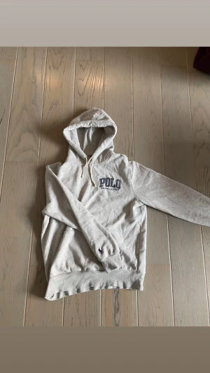 Grå hoodie från Polo Ralph Lauren - Snygg grå hoodie från Polo Ralph Lauren med broderad logga på bröstet och liten häst på ärmen. Tröjan har huva med snörning och ribbade muddar. Perfekt för chill dagar och enkel att matcha med jeans eller joggers.
