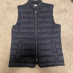 Blå dunväst - Stilren marinblå dunväst med quiltad design och hög krage. Västen har dragkedja framtill och två sidofickor. Perfekt väst nu till våren, säljer då denne för liten för mig tyvärr. Hör av er vid fler frågor eller funderingar!