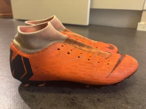 Orange Nike fotbollsskor med strumpkrage - Säljer ett par orangea Nike fotbollsskor med svart logga och strumpkrage för extra stöd runt vristen. Skorna har plös utan snörning och mönstrad ovandel i syntetmaterial. Sulan har fasta dobbar i svart och orange för bra grepp på planen.