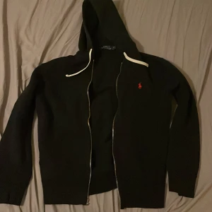 Svart hoodie från Polo Ralph Lauren - Svart hoodie från Polo Ralph Lauren med dragkedja och vit snörning i huvan. Klassisk röd logga broderad på bröstet. Tillverkad i mjuk bomull, perfekt för chill dagar. Långärmad och med en snygg, enkel design.obs dragkedjan e trasig