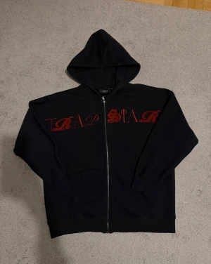 Svart Trapstar hoodie med röd text - Säljer en svart hoodie från Trapstar med dragkedja och stor huva. Framsidan har ett fett rött Trapstar-tryck i unik font. Hoodien har två fickor framtill och ribbade muddar vid ärmar och nederkant. Perfekt för dig som gillar streetwear.