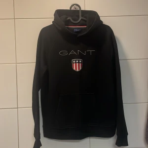 Svart hoodie från GANT med broderad logga - Svart hoodie från GANT med broderad logga och sköld på bröstet. Klassisk känguruficka framtill och huva. Passar dig som gillar stilrena och sportiga plagg med premiumkänsla. Riktigt fint skick. Har använt tröjan kanske 5 gånger och köpte den från Gant helt ny. Riktigt fin hoodie alltså. Passar dig som är mellan 154-164. Nypris är 899kr säljer den för endast 249 riktigt bra pris!