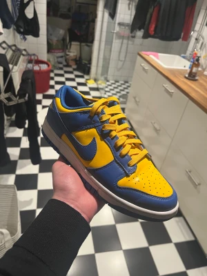 Nike Dunk Low ucla  - Nike Dunk Low sneakers i en fet färgkombo av blått och gult. Skorna har klassisk låg siluett, snörning och rund tå. Ovandelen är i skinn med perforeringar på tån för extra ventilation. Swoosh-loggan i gult på sidan och vit mellansula ger en clean look. Pris kan diskuteras, den köptes i Nike butiken går att bevisa att dem är äkta. Ny pris cirka 2000kr