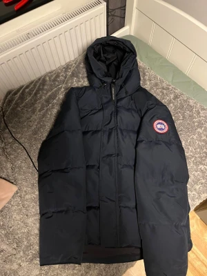 Mörkblå Canada Goose dunjacka - Säljer en mörkblå dunjacka från Canada Goose med huva och klassisk röd-vit logga på ärmen. Jackan har dragkedja framtill och är quiltad för extra värme. Perfekt för kalla vinterdagar och har en clean, oversized look.