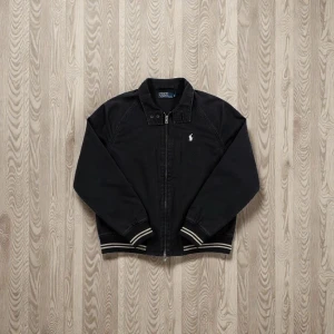 Svart bomberjacka Polo Ralph Lauren - Svart bomberjacka från Polo Ralph Lauren med klassisk krage, dragkedja framtill och vit broderad logga på bröstet. Jackan har ribbade muddar med vita ränder och är tillverkad i bomull. Perfekt för dig som vill ha en clean och tidlös look. 🙌  Skick 7.5/10 ✅ Inget fel på den. Snabb leverans 🚚