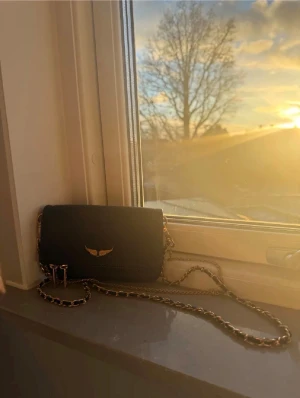 Zadig voltaire väska - Snygg svart axelväska i skinnimitation med guldfärgad kedja och ett elegant guldfärgat emblem framtill. Väskan har dragkedja upptill och stilren design, perfekt för att lyfta din outfit. Kedjan är flätad med svart material för extra detalj.