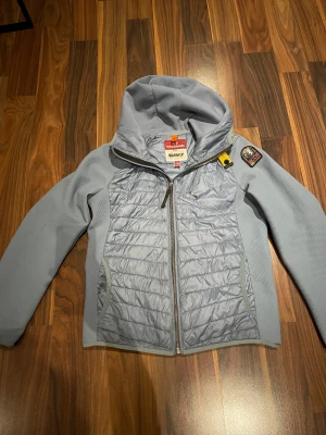Ljusblå Parajumpers pufferjacka med huva - Snygg ljusblå pufferjacka från Parajumpers med huva och coolt PJS-märke på ärmen. Jackan har quiltad framsida och rygg, dragkedja framtill och praktiska fickor. Materialmix med polyester och syntet, perfekt för dig som gillar sportig stil. Ny pris 5000kr