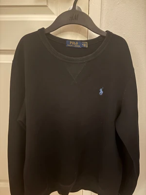 Svart sweatshirt från Polo Ralph Lauren - Svart sweatshirt från Polo Ralph Lauren med klassisk rund hals och blå broderad logga på bröstet. Tröjan har långärmad passform och är tillverkad i mjuk bomull, perfekt för chill dagar. Snygg och enkel design som funkar till jeans eller joggers.