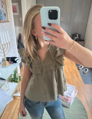 Grönt blus med volanger från Gina Tricot - Supersöt beige blus från Gina Tricot med volangdetaljer på axlarna och runt bröstet. Blusen har v-ringning med knyt framtill och är ärmlös. Luftig och flowy passform, perfekt för vår och sommar. Materialet är mjuk bomull som känns skönt mot huden.