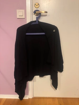 Svart omlottkofta med knappdetalj - Svart kofta med omlott-design och en snygg knappdetalj på axeln. Koftan har långa ärmar och ett draperat fall som ger en chill och modern vibe. Perfekt att slänga över en t-shirt eller linne när det är lite kyligt ute.