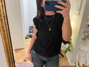 Svart blus med volang från Carin Wester - Svart blus från Carin Wester med volangdetaljer längs axlarna och ärmlös design. Blusen har en lätt textur i tyget och rund halsringning. 