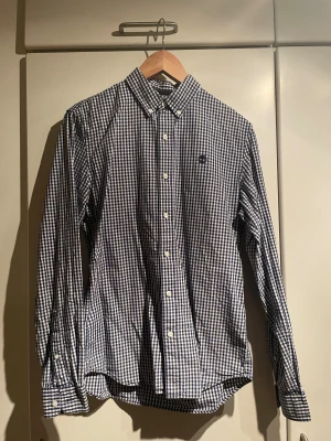 Blårutig skjorta från Timberland - Snygg blårutig skjorta från Timberland i slim fit-modell. Skjortan har klassisk krage, vita knappar och lång ärm. Materialet är mjuk bomull och mönstret är små rutor i blått och vitt. Perfekt för dig som gillar stilrena och tidlösa plagg.