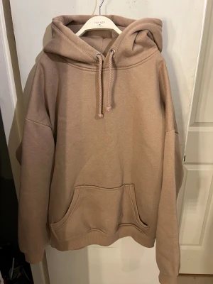 Beige hoodie med huva och ficka - Säljer en beige hoodie från Bik Bok i storlek S. Tröjan har en stor huva med snörning och en klassisk magficka framtill. Modellen är loose fit med långa ärmar och ribbade muddar.