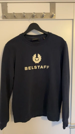 Mörkblå sweatshirt - Belstaff - Mörkblå sweatshirt från Belstaff med vitt logotryck på bröstet. Tröjan har rund hals, långa ärmar och är gjord av bomull. Stabil kvalité. Storlek M men sitter som en S eller en mindre M. Nypris 2299kr. Använd väldigt få gånger då jag växt ur den och skicket är som nytt. Fått i present därav finns inte kvitto, men kan naturligtvis skicka bilder på tags osv. Hör av er för specifika bilder så ordnar jag det. 